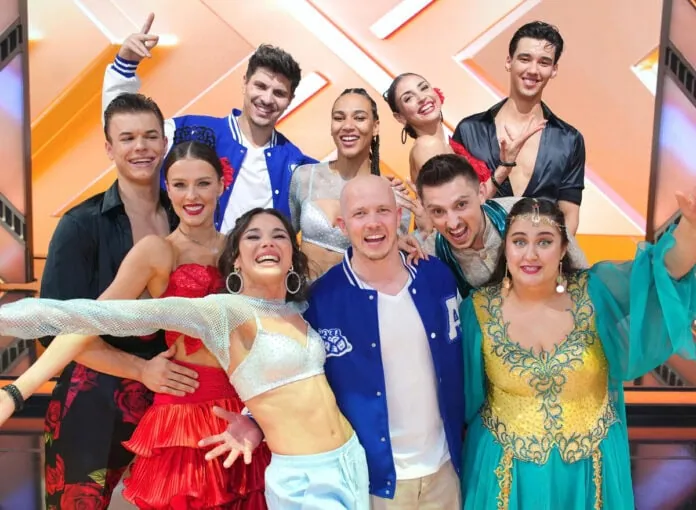 Cast von Let's Dance in bunten Bühnenkostümen mit glitzernden Accessoires vor orangefarbenem Bühnenhintergrund