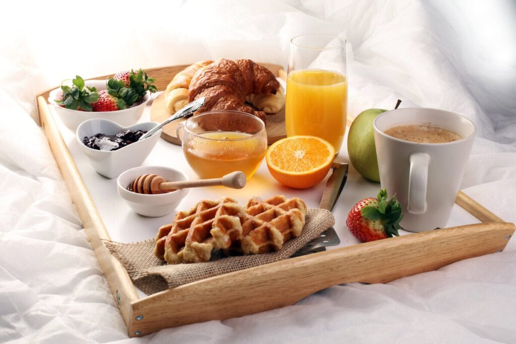 Liebevoll vorbereitetes Frühstück im Bett auf Holztablett mit Croissant, Obst, Orangensaft und Kaffee auf weißem Bettzeug
