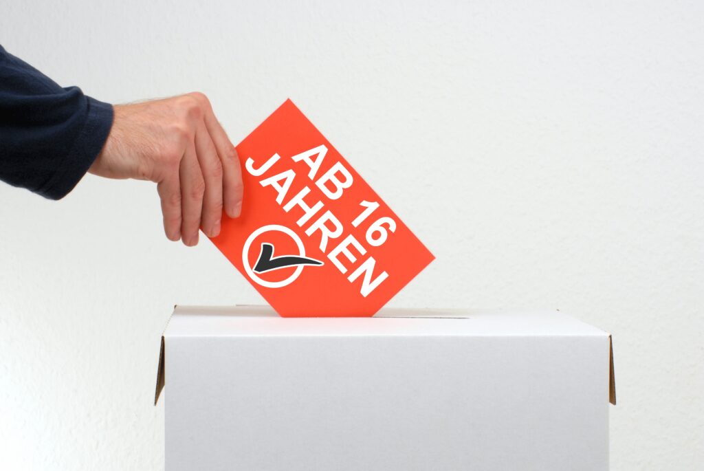 Stimmzettel mit Aufschrift Ab 16 Jahren wird in die Wahlurne geworfen